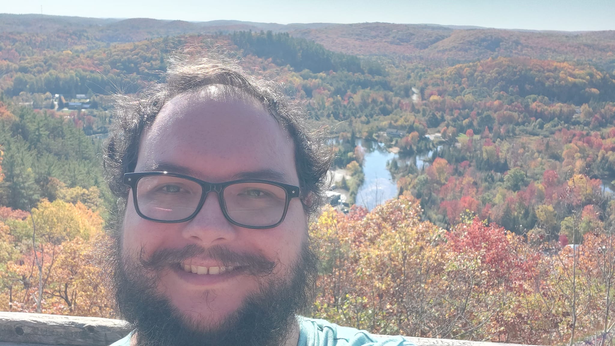 A man with short hair and bushy facial hair stands in front of a wooden fence seperating him from a steep hill covered in autumn colours, hills stretch out to the horizon, some covered in green conifers, others the red and gold leaves of the season, a river snakes through the midground